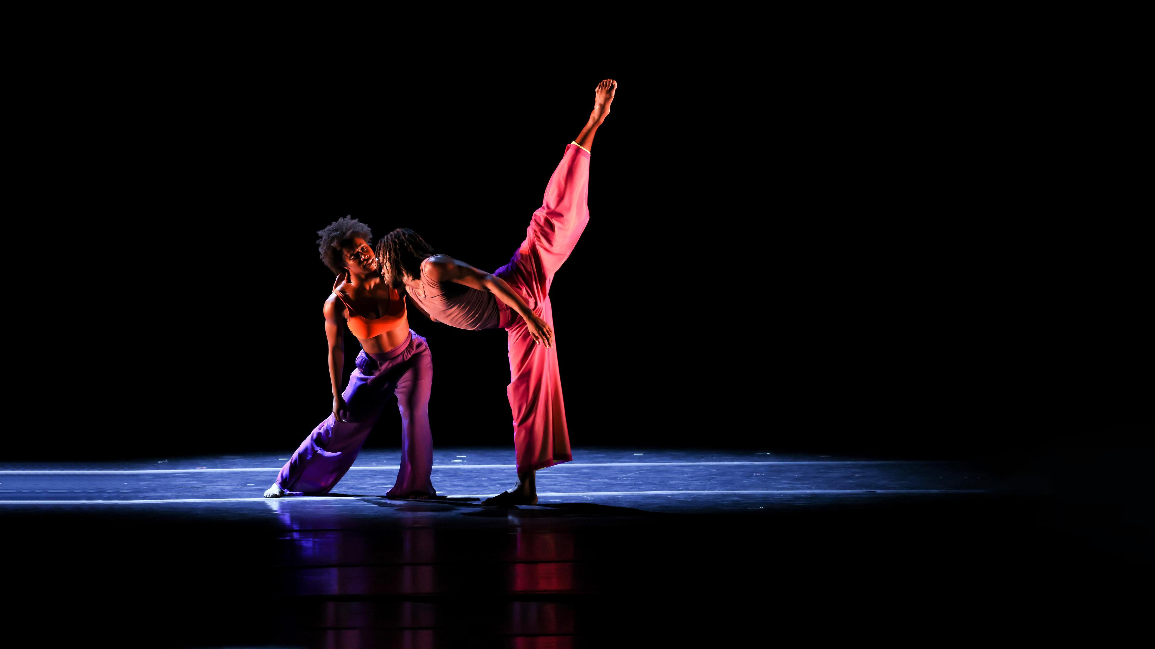 A woman in an orange top and purple pants leans next to a man in a pink top and pants leaning forward with one leg in the air.