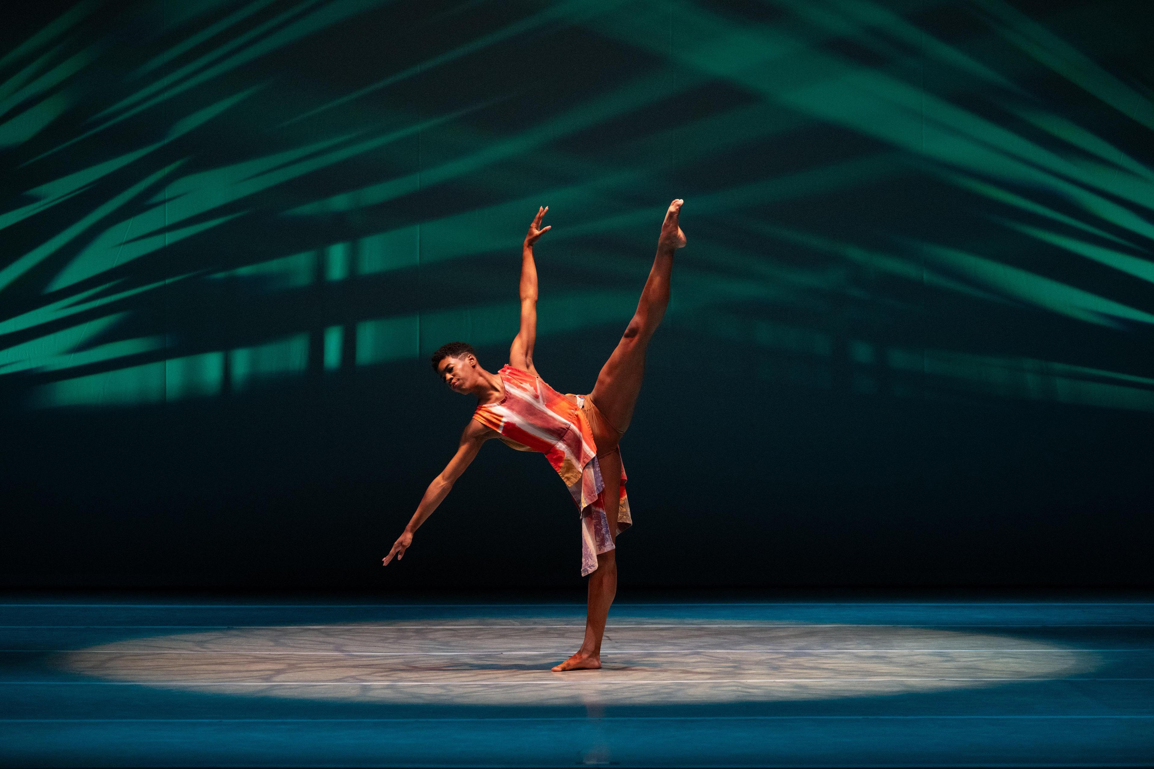 A woman in an orange dress tilts to the side with one leg in the air and head looking towards the ground.