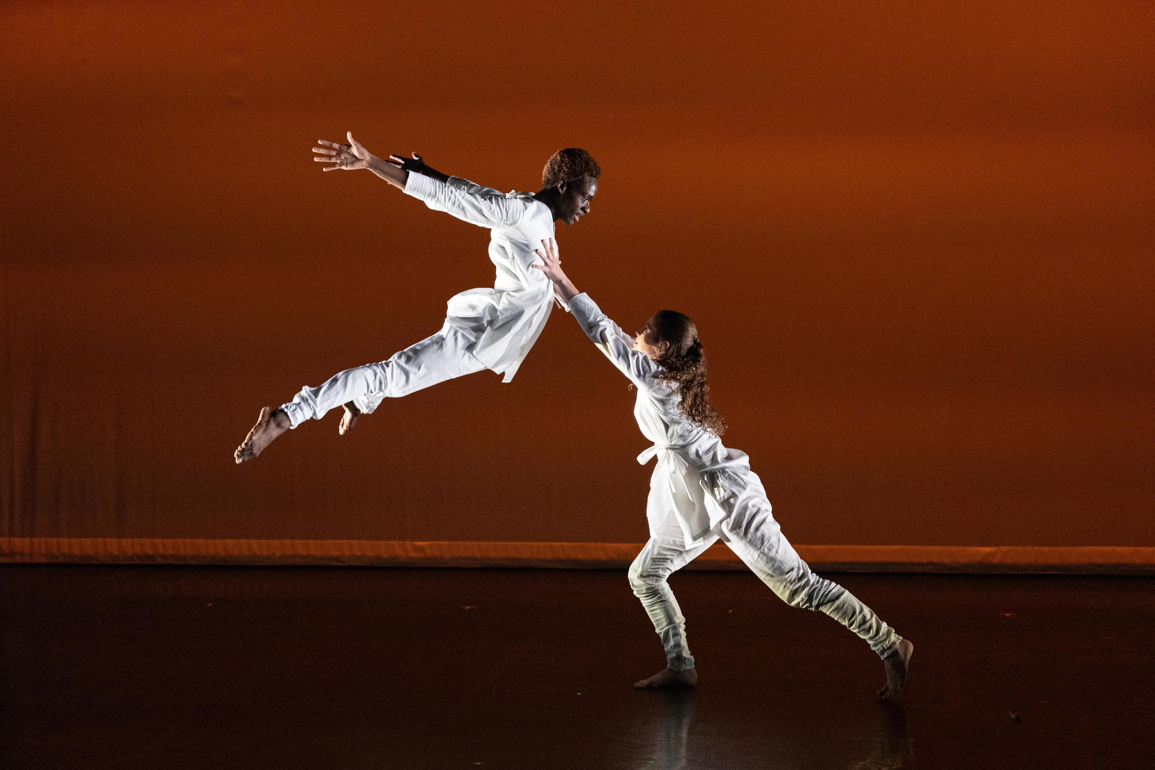 Two dancers dressed in white, on jumping up in the air towards the second one standing nearby