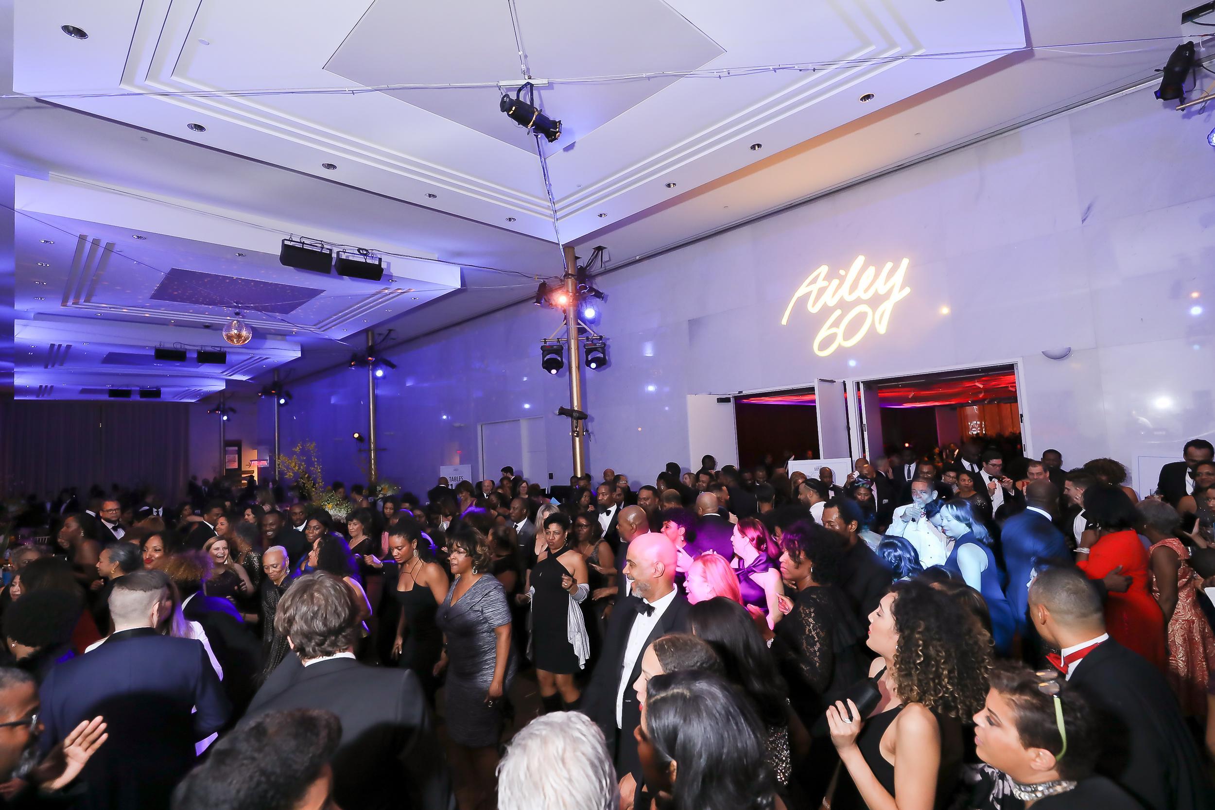 Ballroom full of people dancing. There is an LED sign on the wall that says "Ailey 60". The room has multicolored party lights.