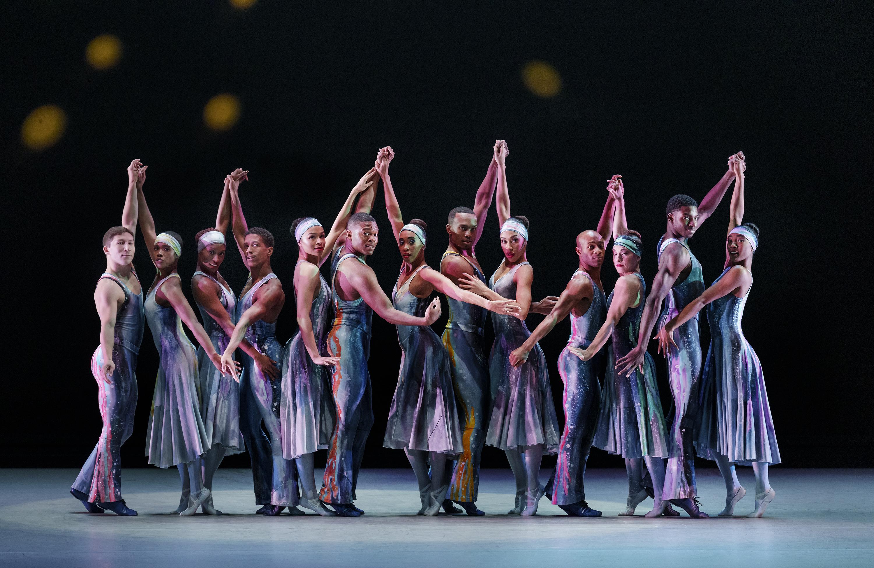 A group of dancers stand together in a line wearing flowy tie dye costumes