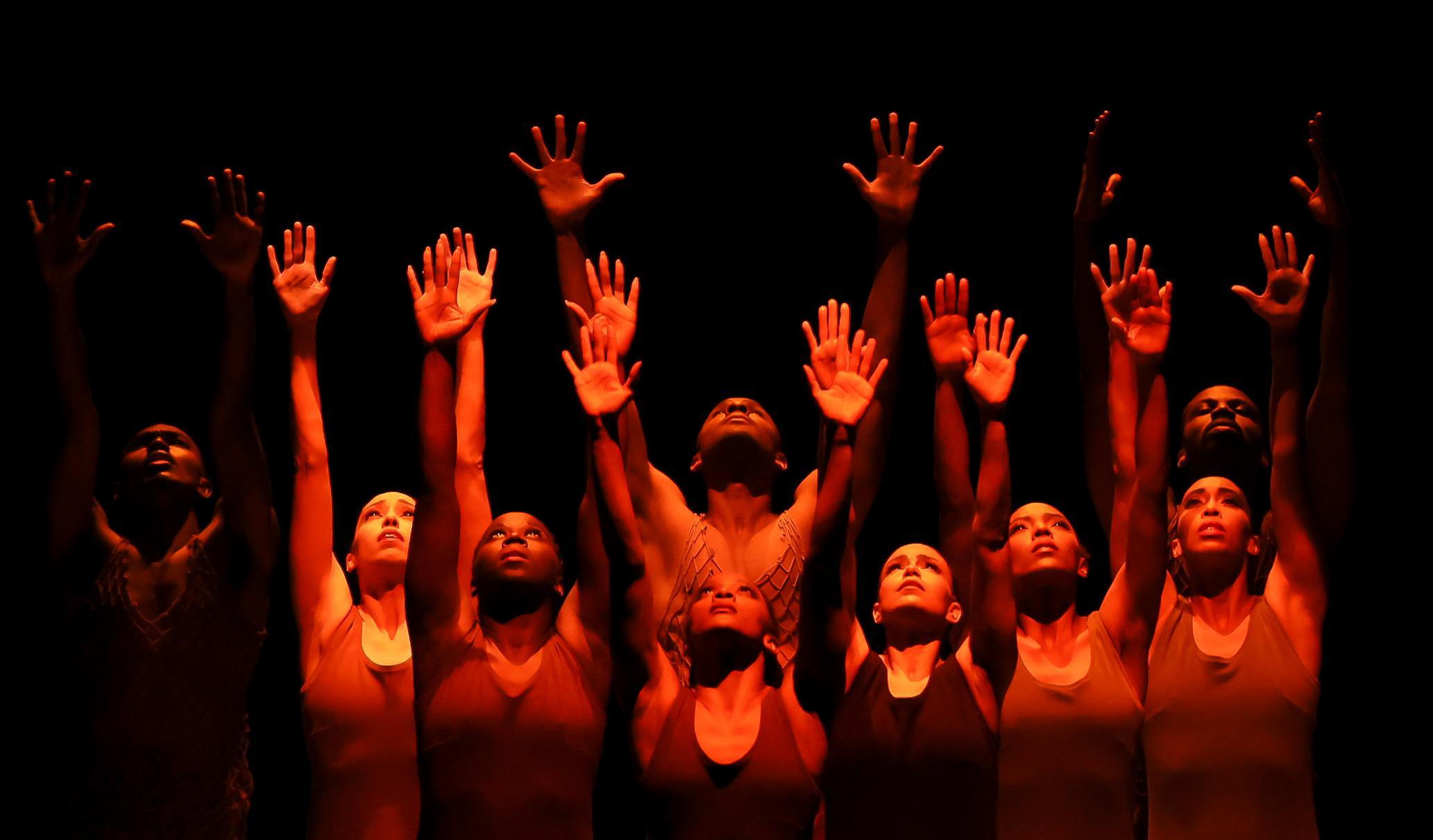 A group of dancers in flesh tone dresses look up and reach their hands to the sky 