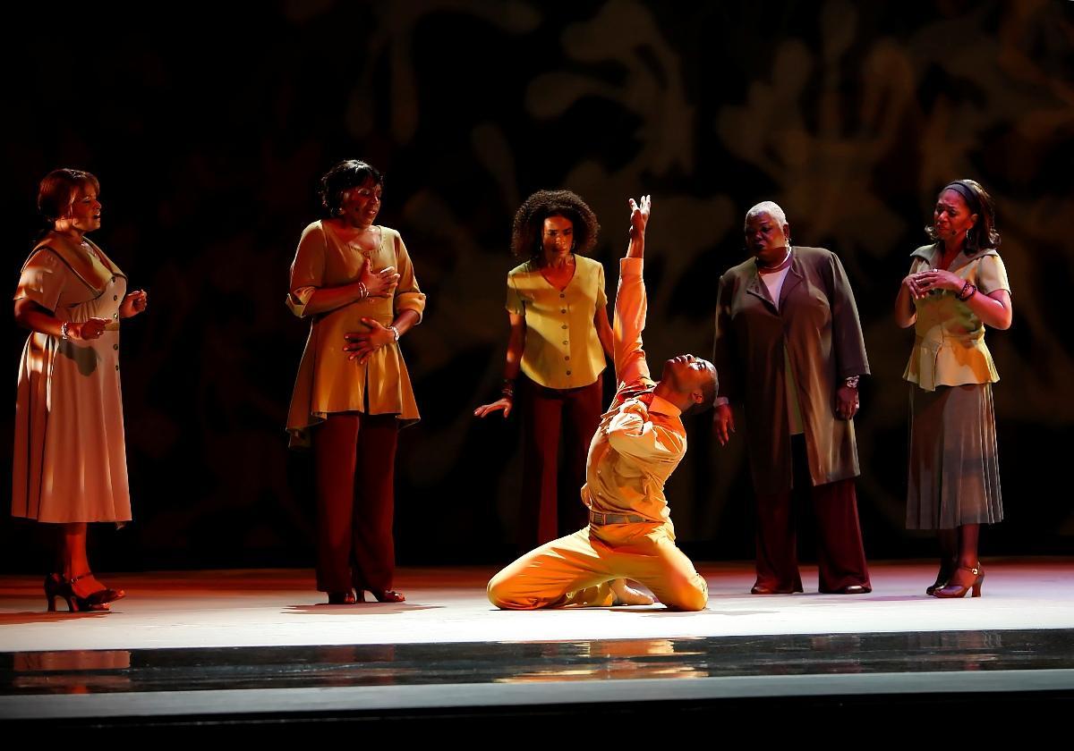 A group of people stand on stage and one sits on the ground reaching arms up in the middle of the stage