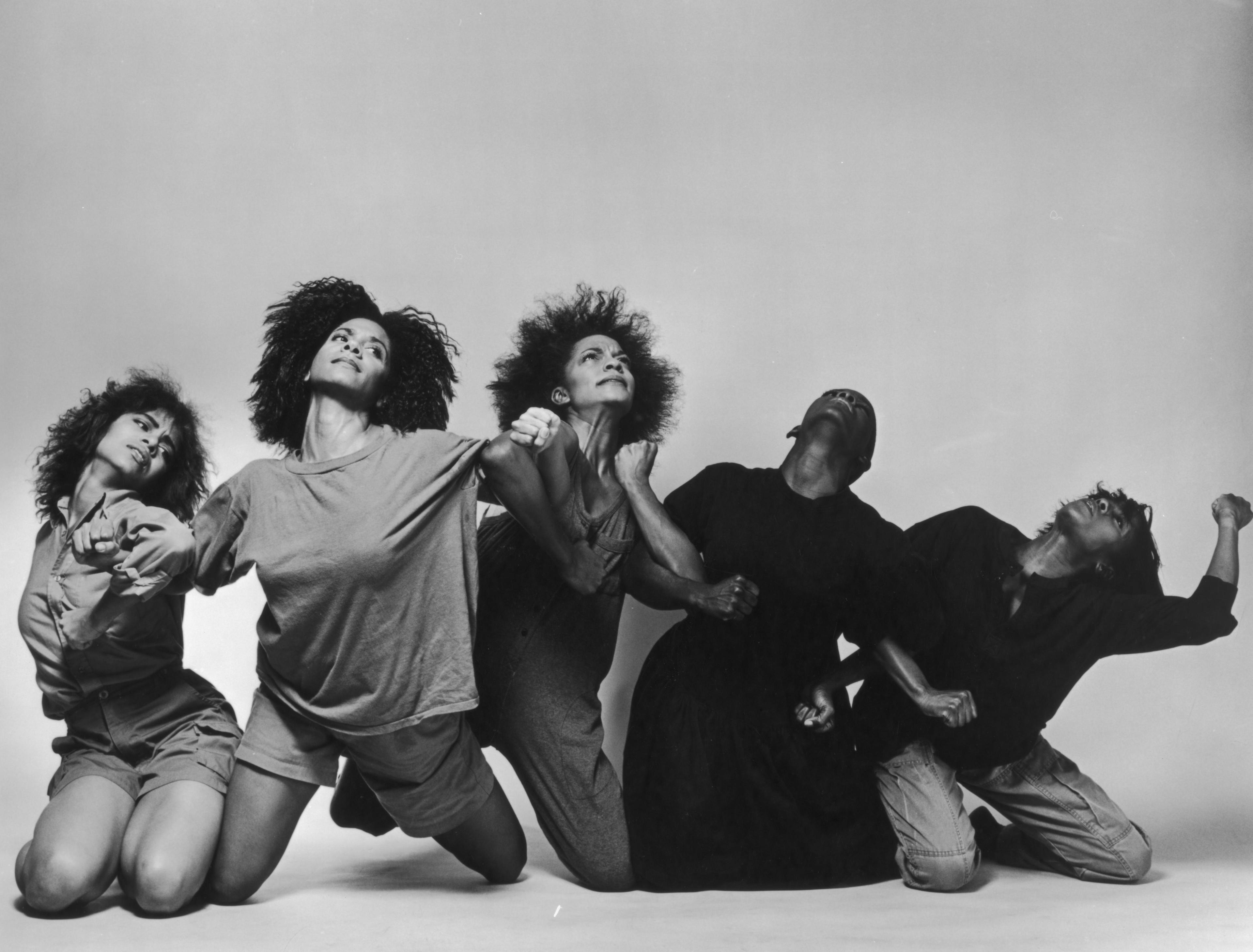 five women sit on the ground arms linked looking up at the sky in anquish.