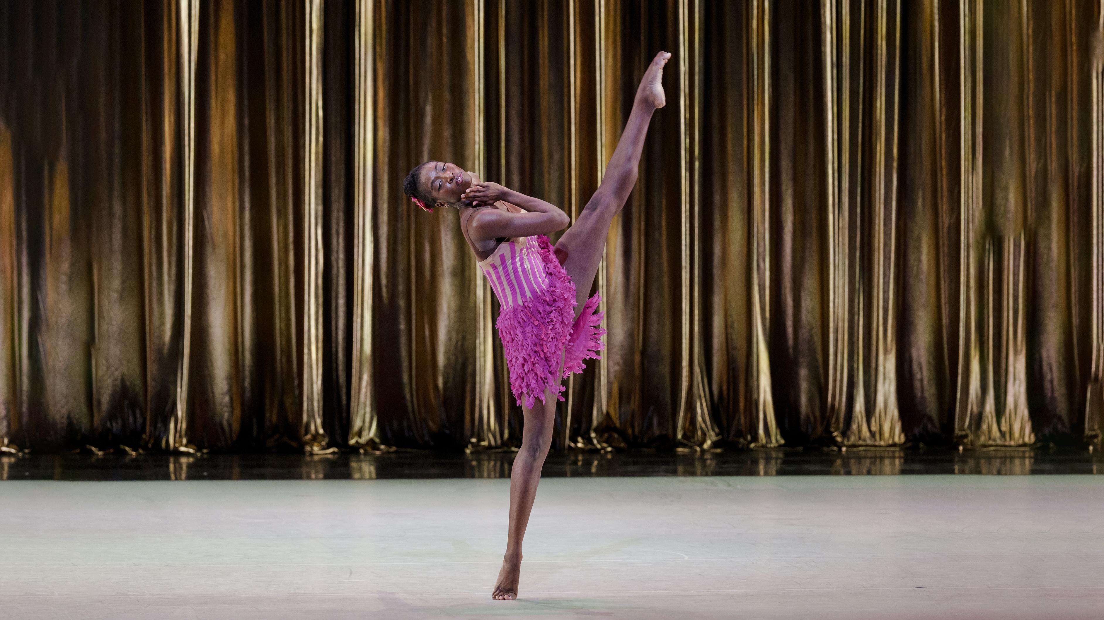 A woman in a pink dress kicks one leg up into the air.