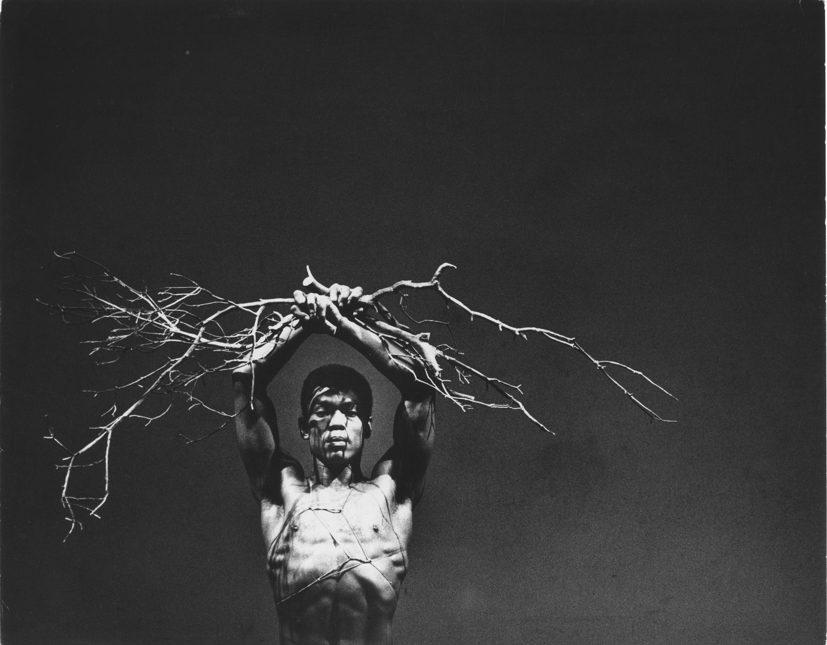 A shirtless man holds tree branches over his head