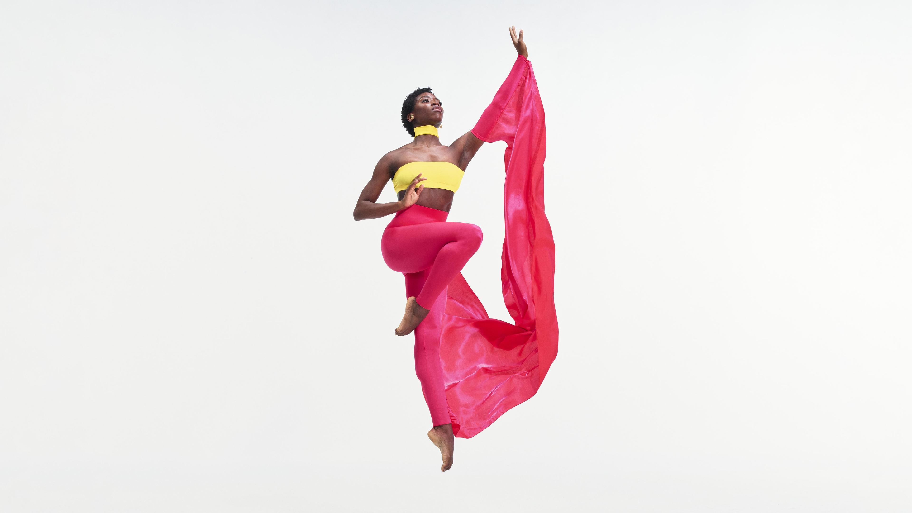 A female dancer wearing a yellow tube top and pink pants jumps in the air holding a swath of pink fabric