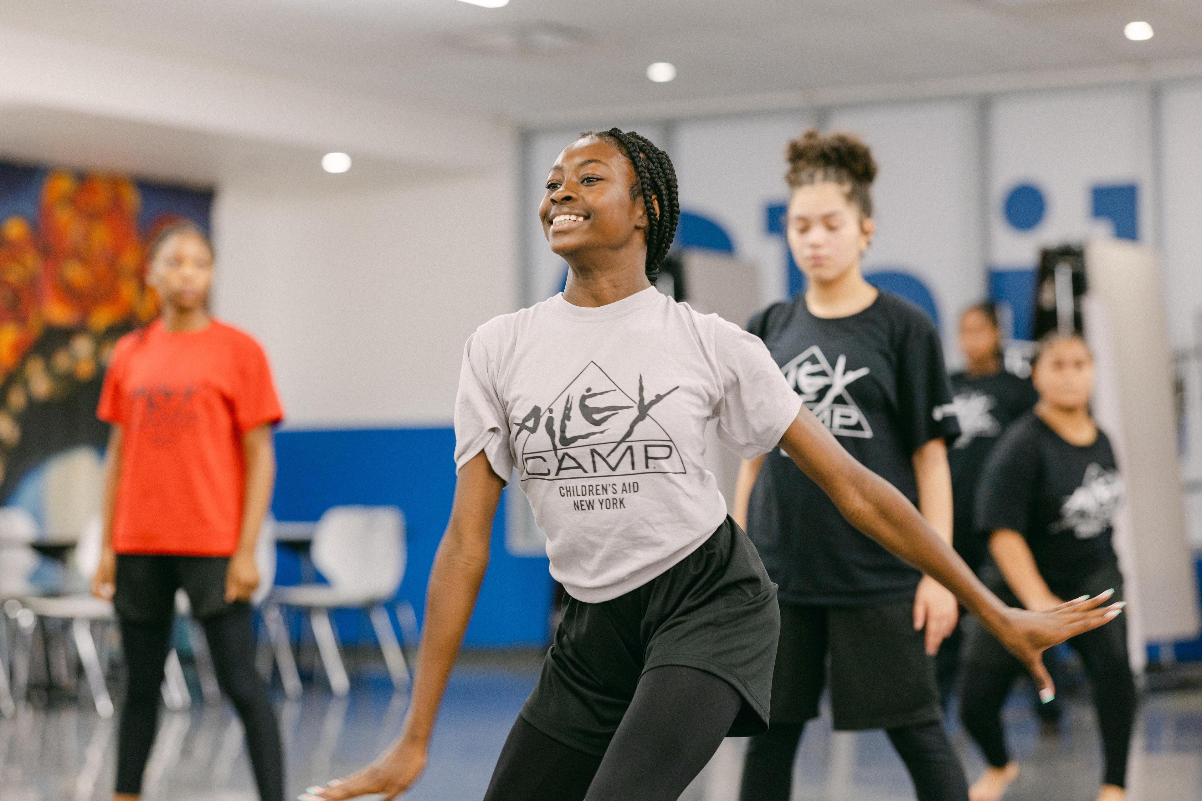 A girl in a gray t-shirt and black shorts smiles while dancing in a studio with other children.