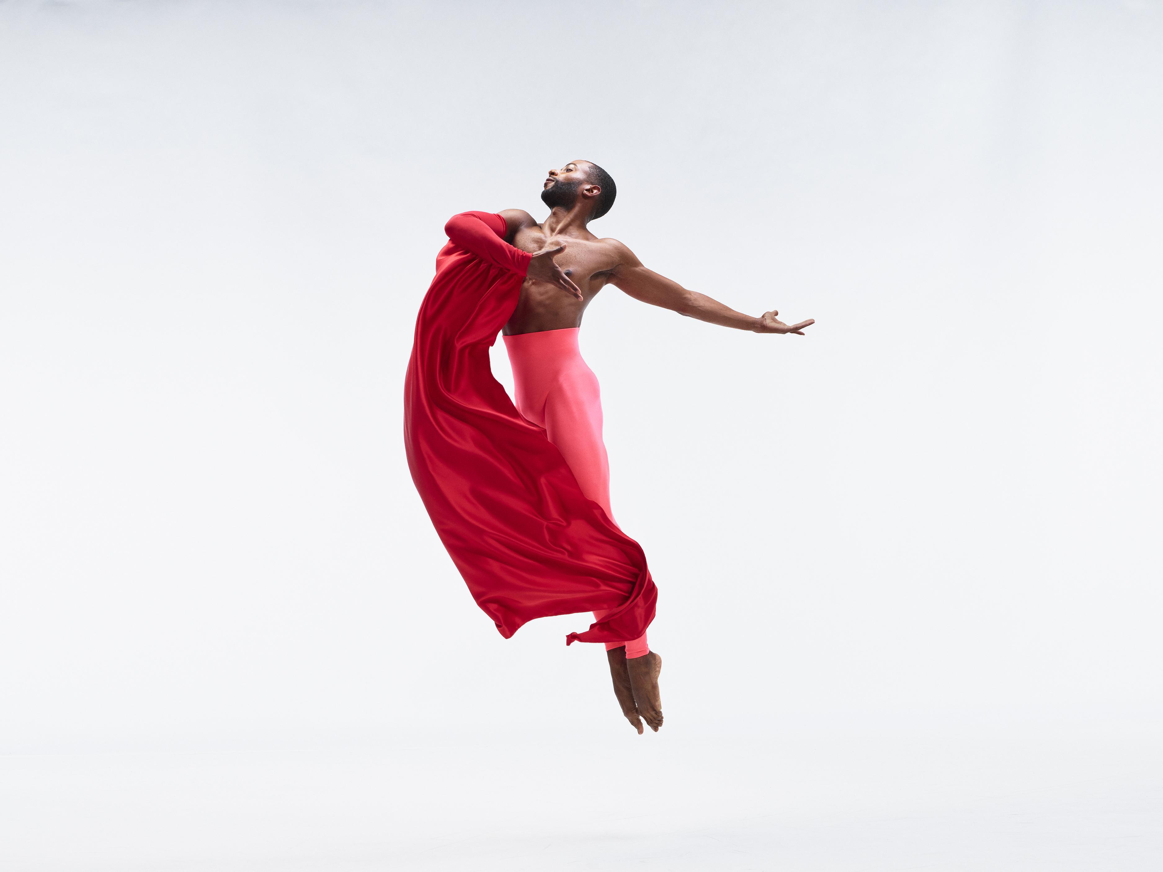 A shirtless man wearing bright pink pants and holding a swath of red fabric jumps up into the aair