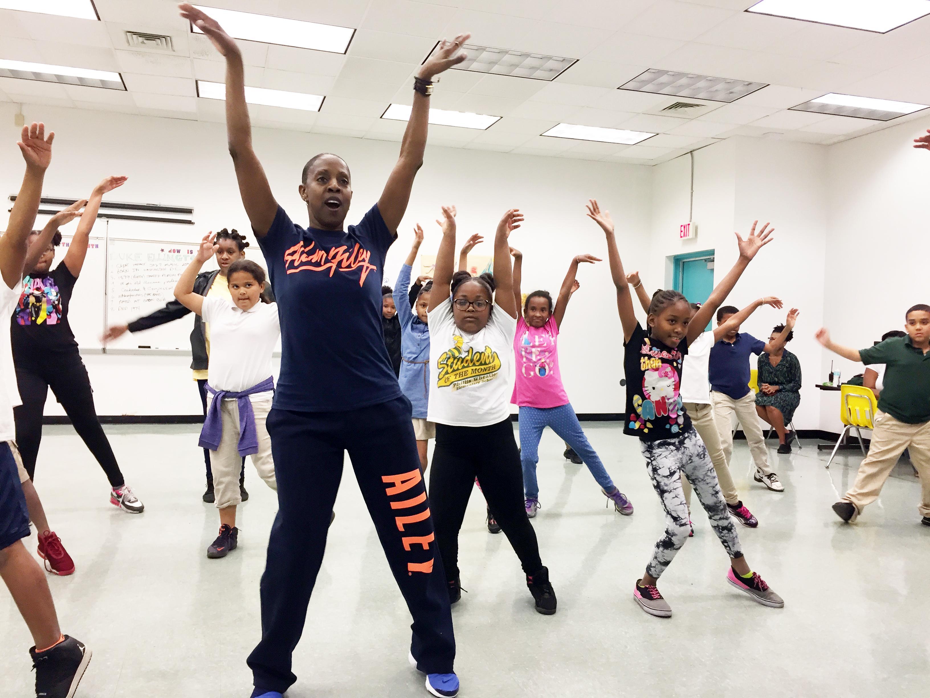 A group of students moving in a dance studio with their arms raised in the air.