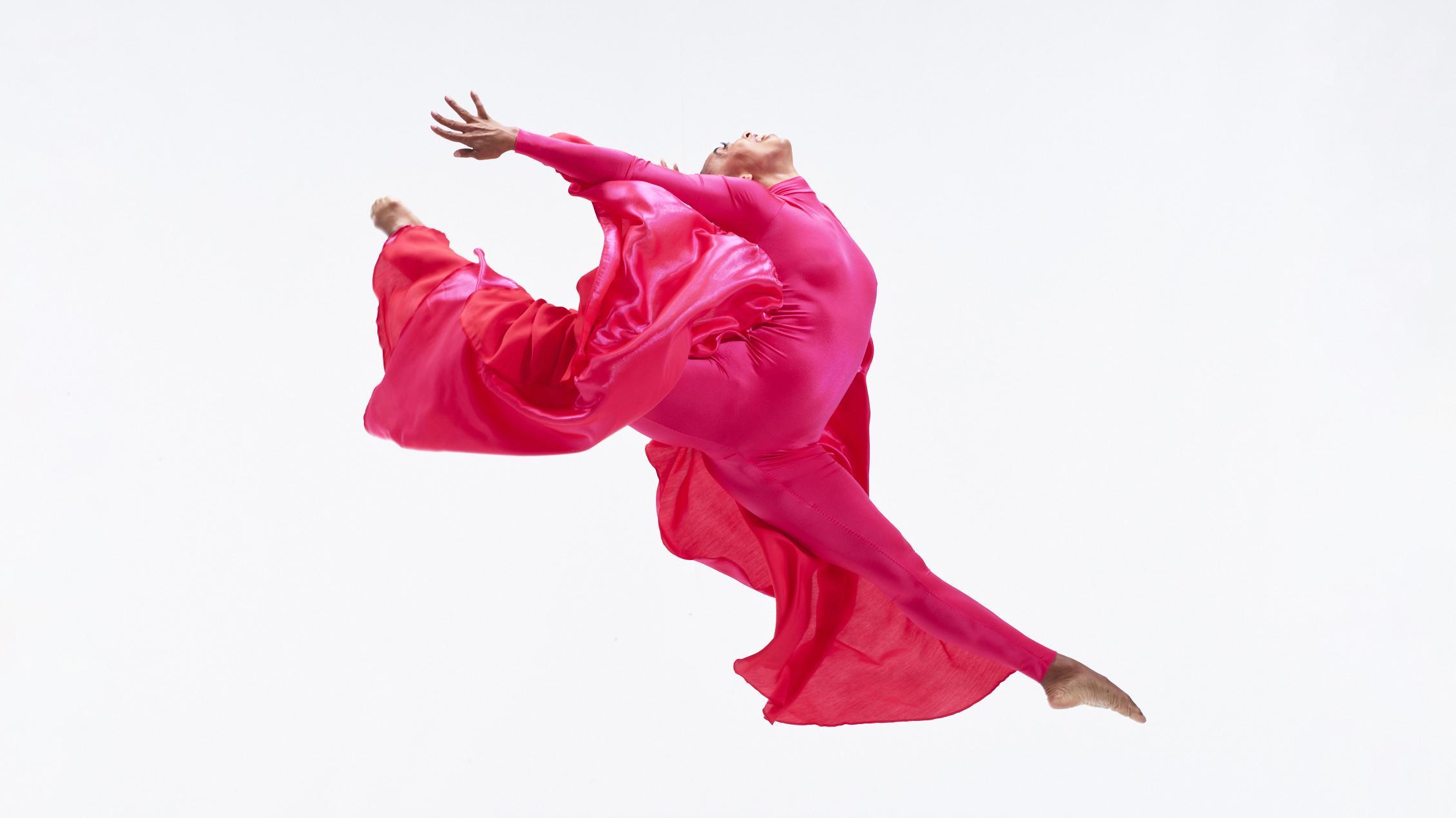 A dancer in a hot pink ensemble. They are jumping mid-air with their hands above their head.