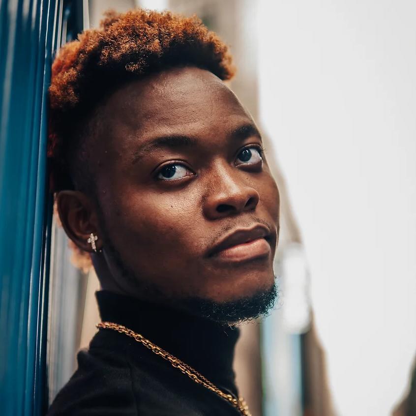 Headshot of a black man with short dark hair with red on the tips, wearing a black turtleneck with a gold chain and earrings