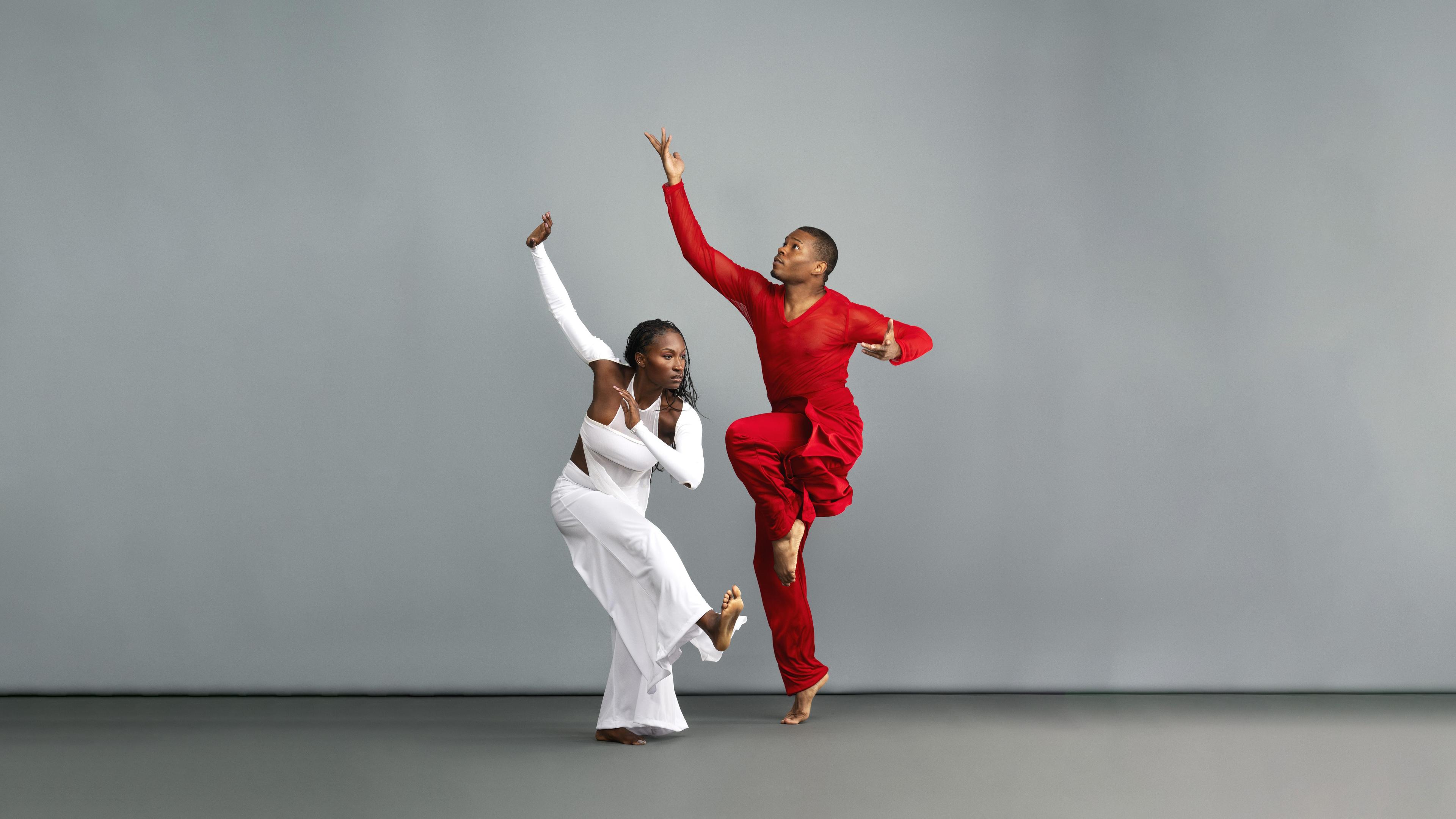 Two dancers, one in red and one in blue. They are dancing in a grey studio.