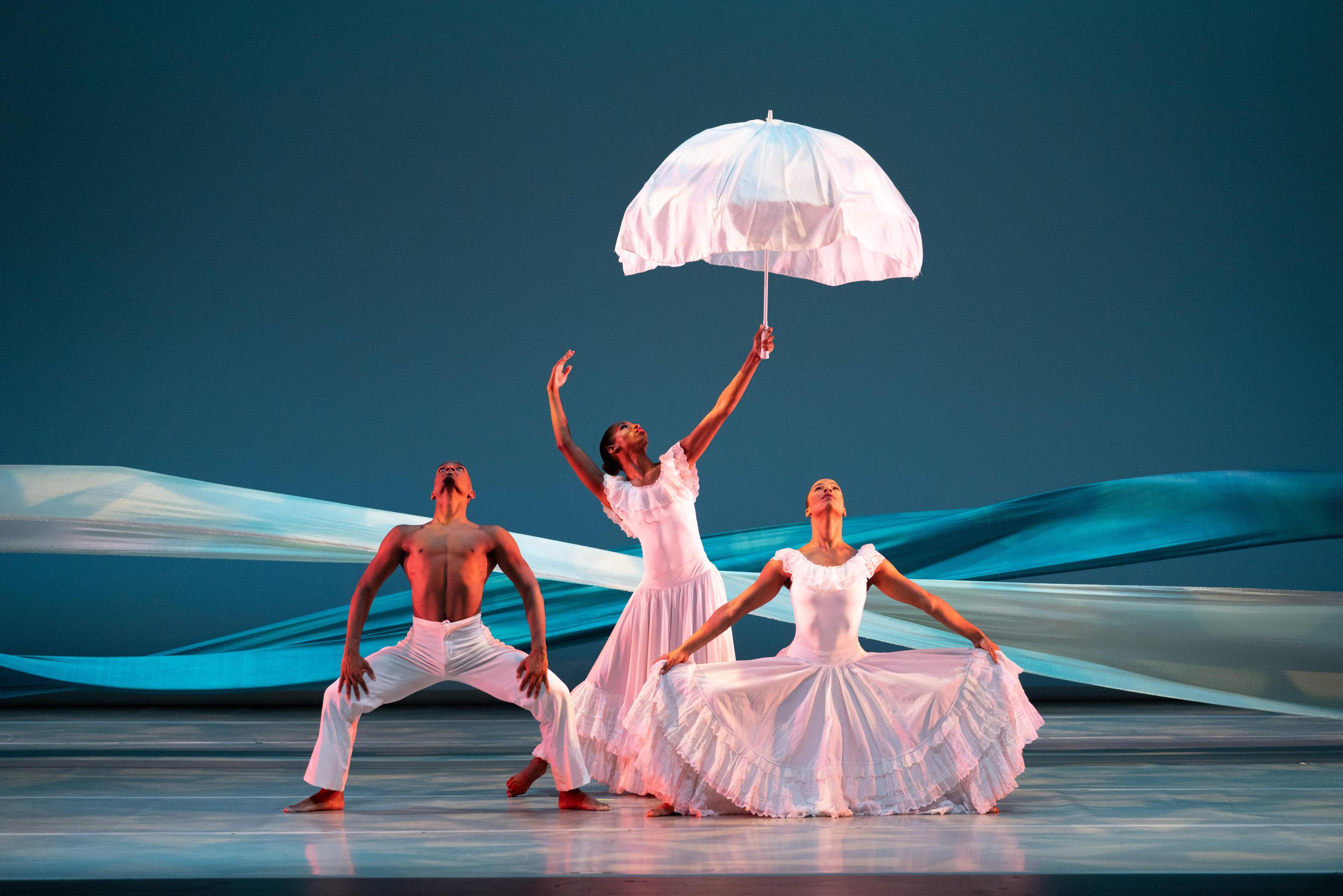 3 dancers in white, 1 holding an umbrella and white fabric billowing behind them with blue lighting looks like water