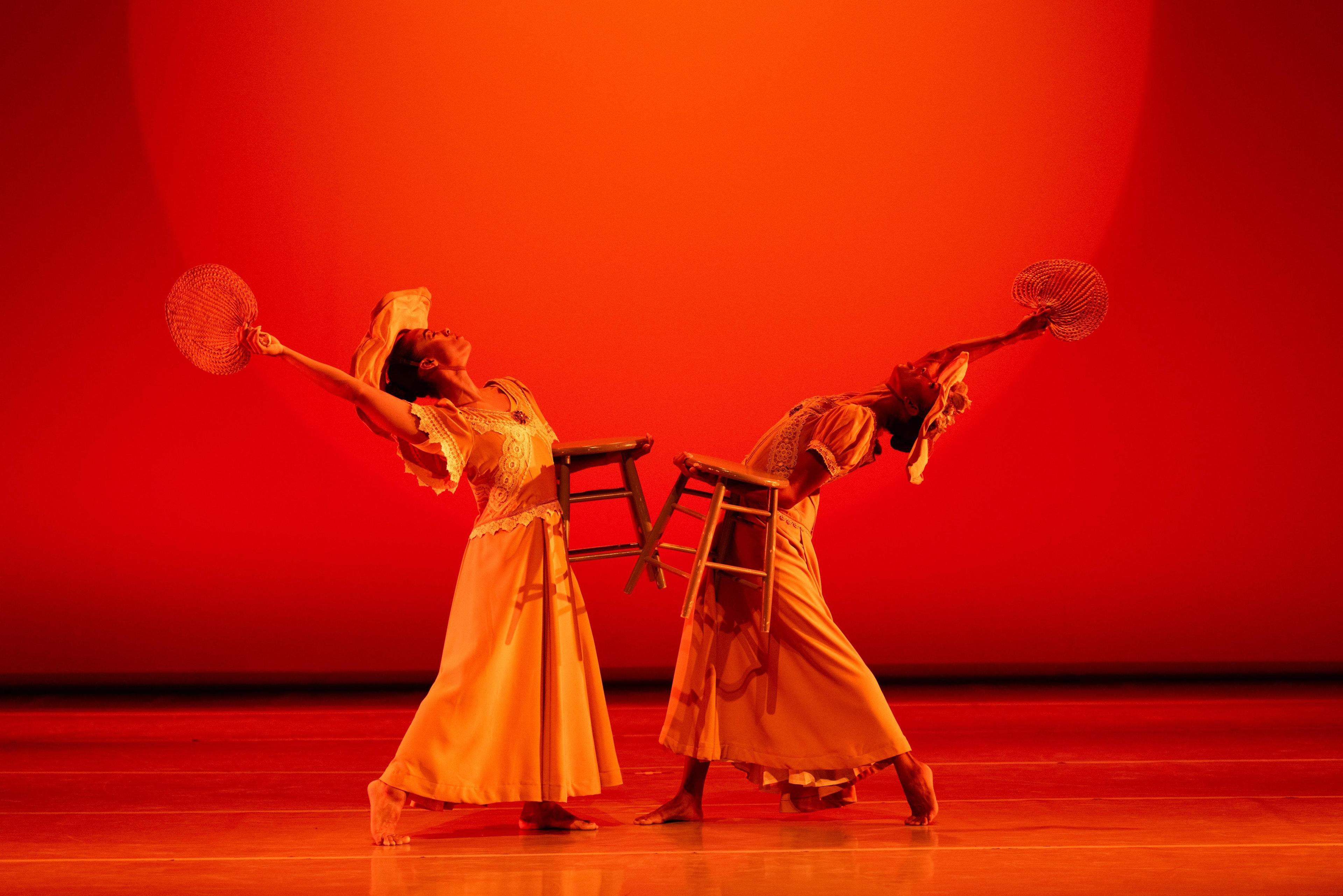 2 dancers in yellow arched away from each other holding a fan and a stool