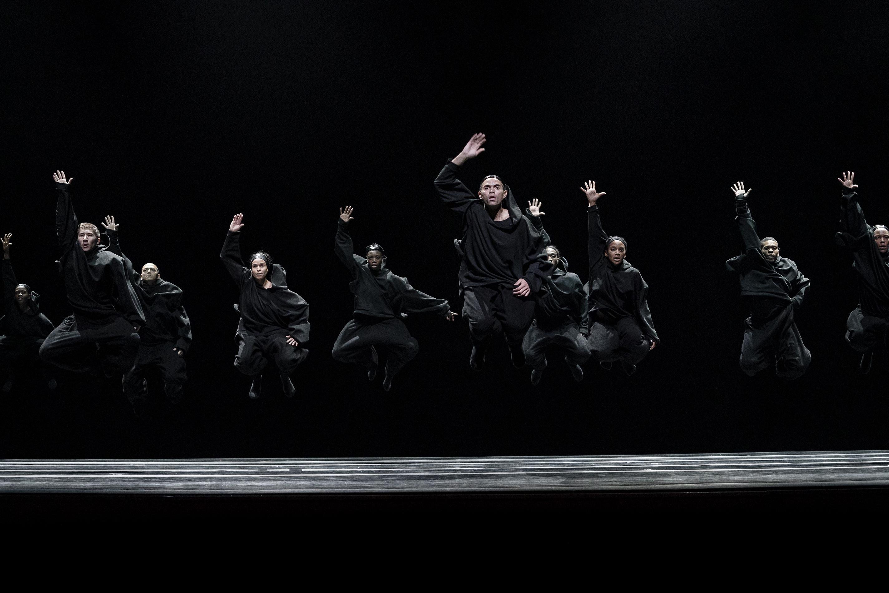 A group of dancers dressed in black hoodies and black pants jumps up into the air with one hand raised above their heads.