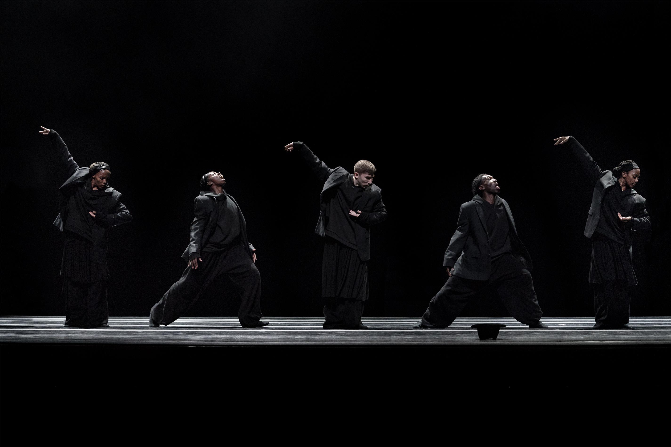 Five dancers lined up side by side. Every other dancer is raising their arms backwards and holding their other arm in front of them while the other dancers extent their chests and look up