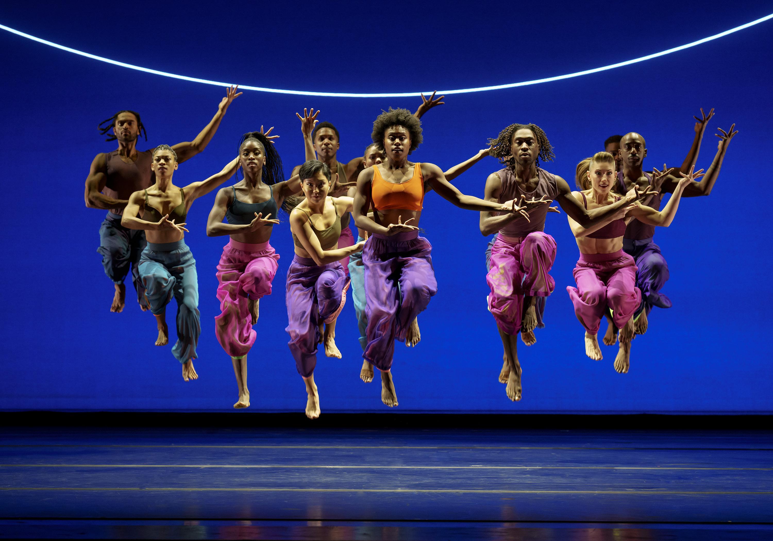 A group of dancers from the Alvin Ailey American Dance Theater performing "Are You in Your Feelings" by Kyle Abraham. They are captured in mid-jump, wearing colorful flowing pants and crop tops, set against a bright blue background with a curved light above them.