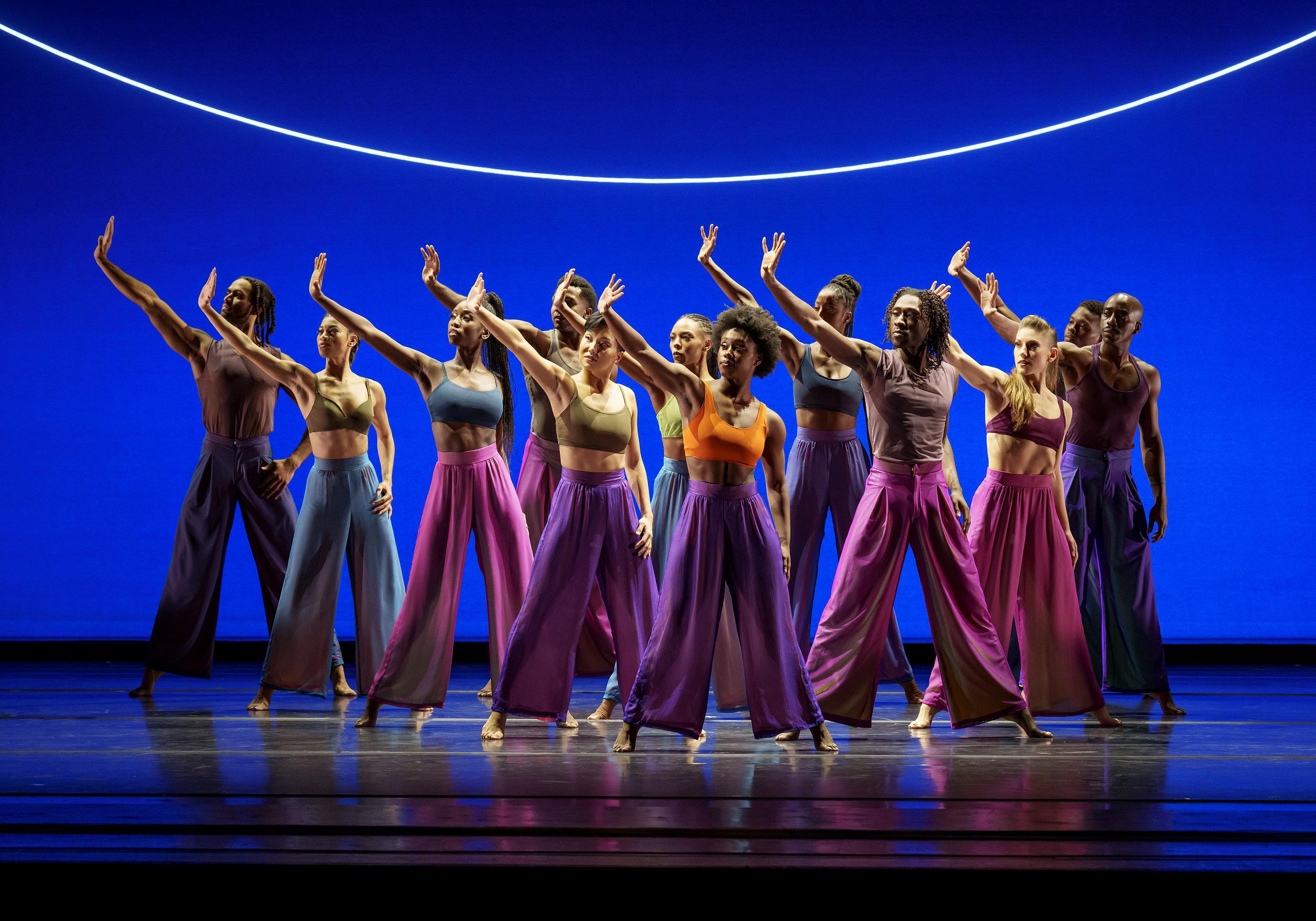 A group of dancers from the Alvin Ailey American Dance Theater performing "Are You in Your Feelings" by Kyle Abraham. They are on stage in vibrant, flowing pants and crop tops, with a blue background and a curved light above them.