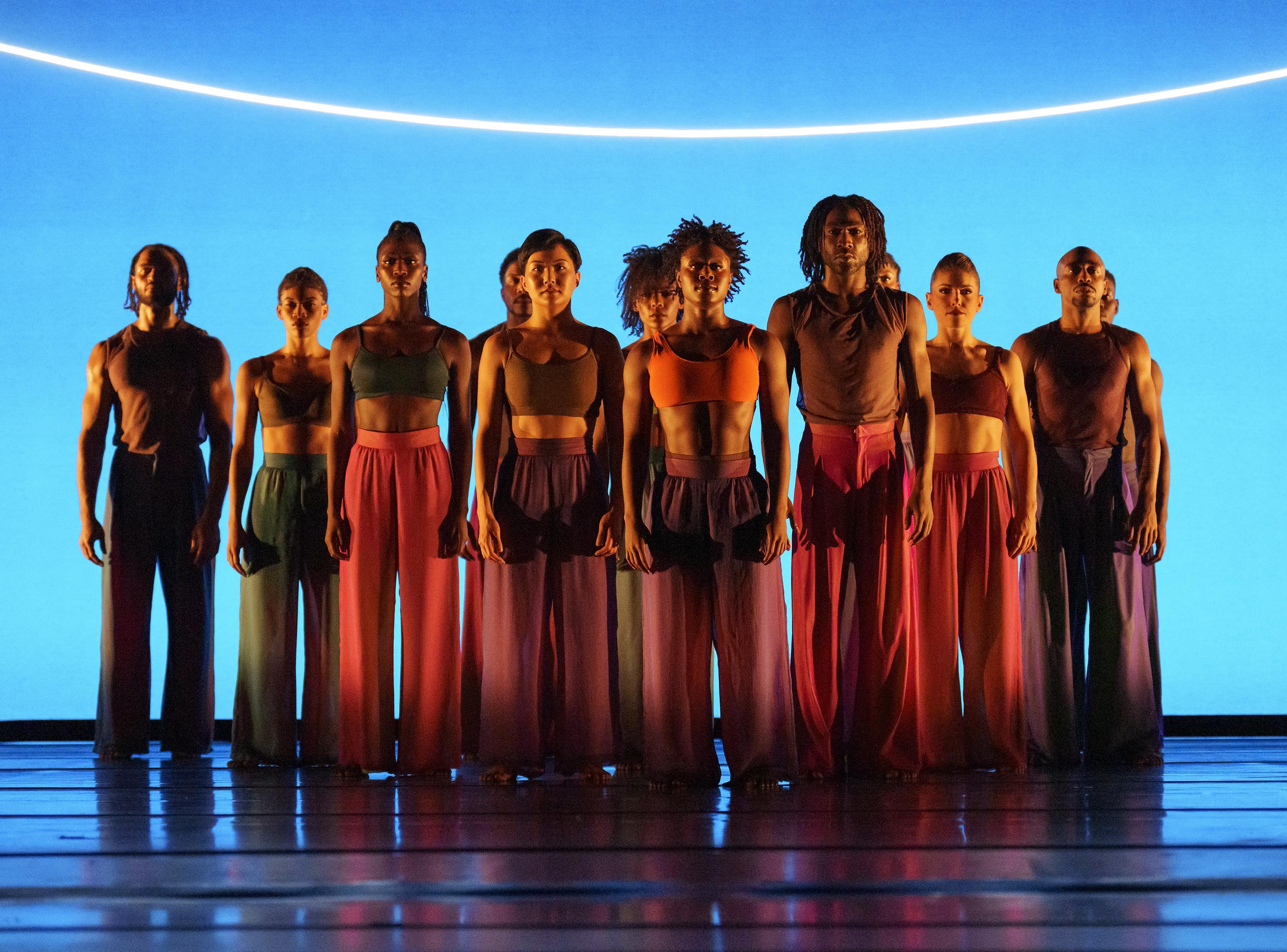 A group of dancers from the Alvin Ailey American Dance Theater standing in a line, performing "Are You in Your Feelings" by Kyle Abraham. They wear vibrant, flowing pants and crop tops against a bright blue background with a curved light above them.