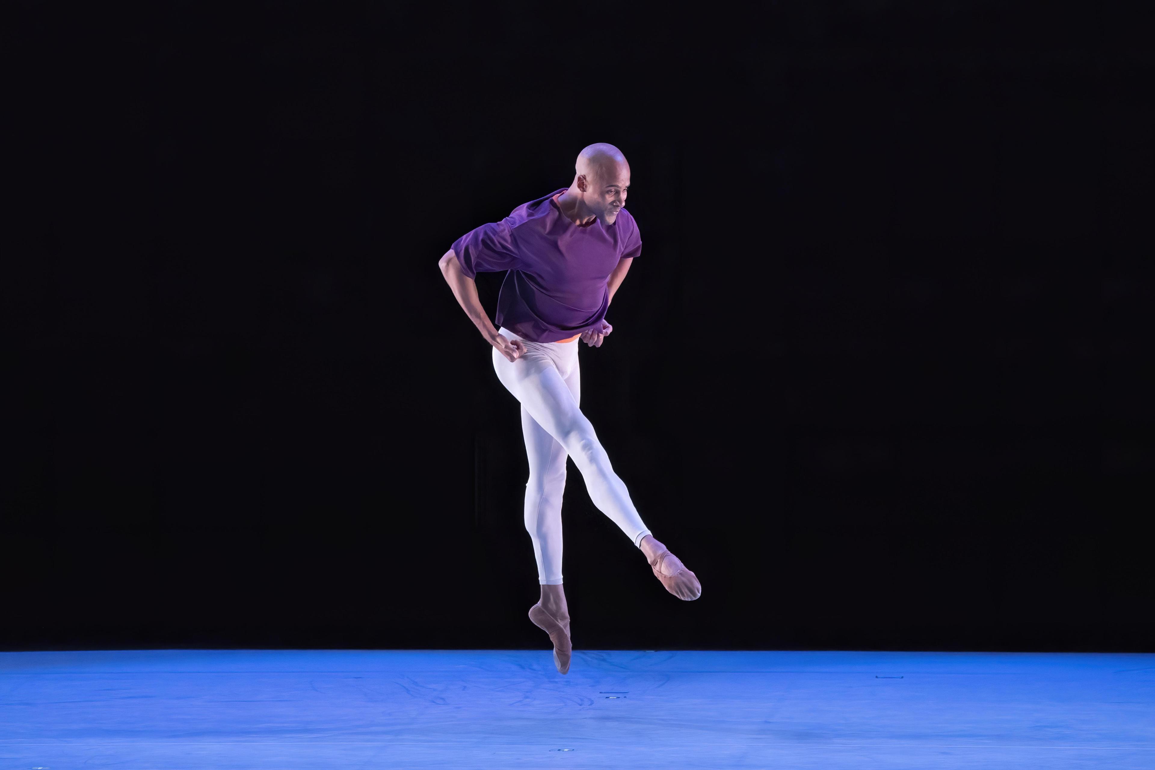 Dancer mid-jump. They are wearing a purple shirt and white pants. They are alone on stage.