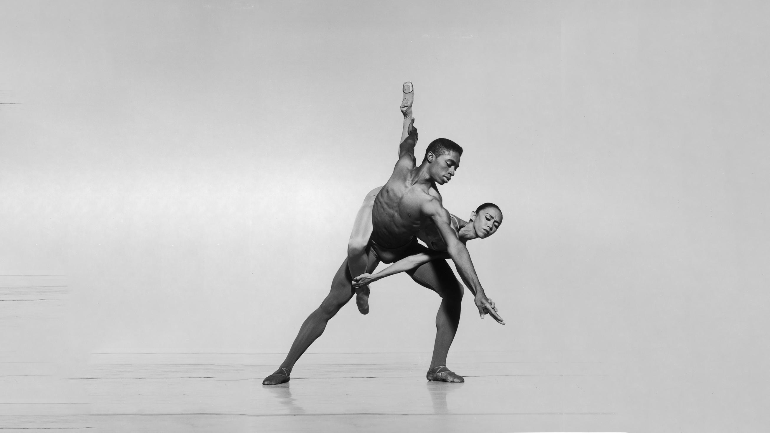 A black and white photo of two dancers, Desmond Richardson and Elizabeth Roxas, in mid-performance. Desmond Richardson is shirtless, leaning forward with one arm extended, while Elizabeth Roxas is lifted behind him, one leg extended upward. They both wear ballet shoes, and the background is a plain light color. The photo is labeled REPERTORY Desmond Richardson and Elizabeth Roxas in After Eden. Photo by Jack Mitchell.