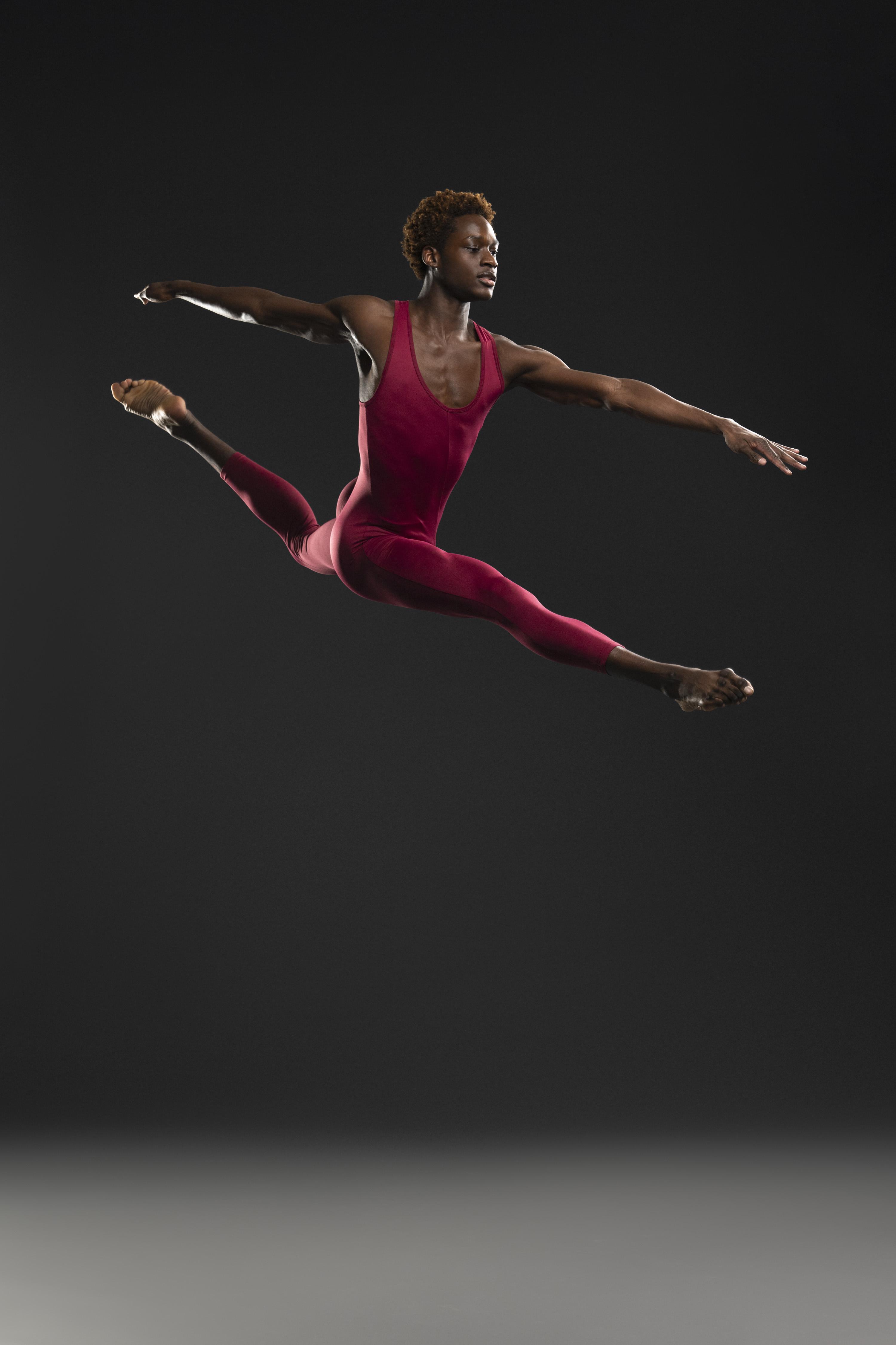 A man in a burgundy unitard jumps in the air with legs in an attitude split