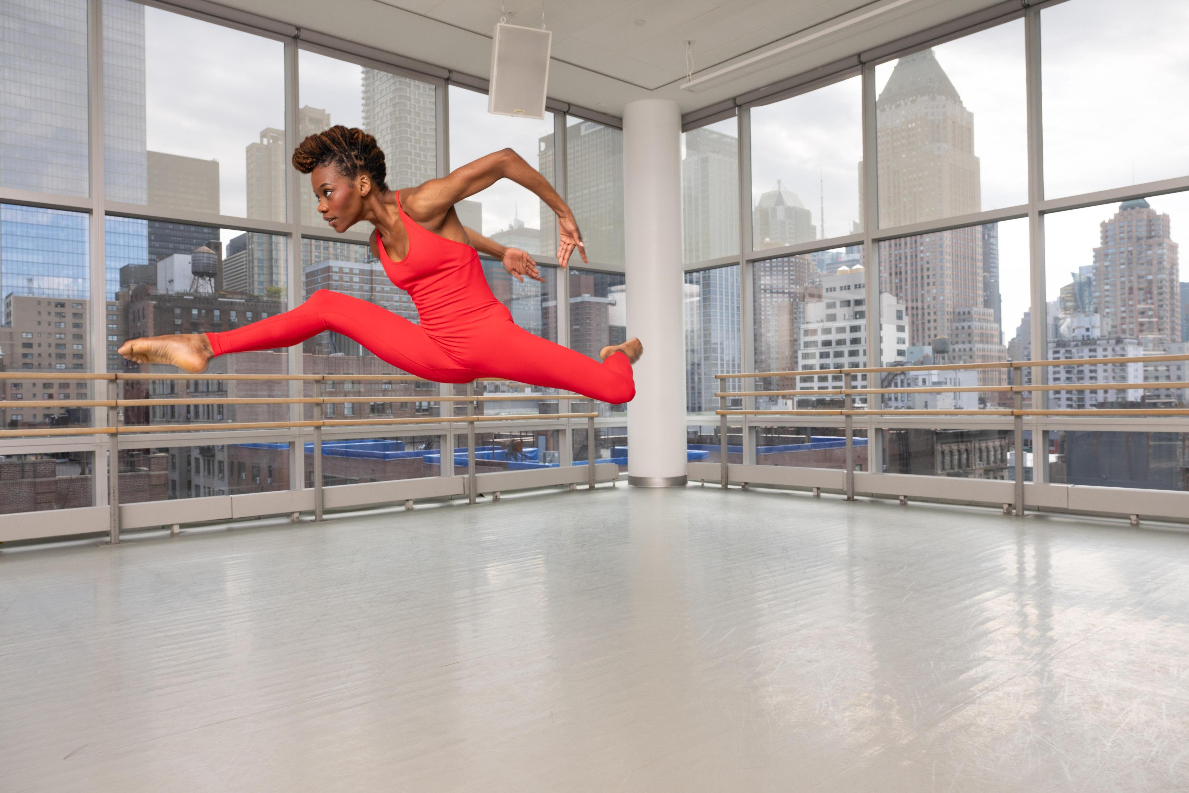 A woman in a bright orange unitard jumps in the air with legs in an attitude split